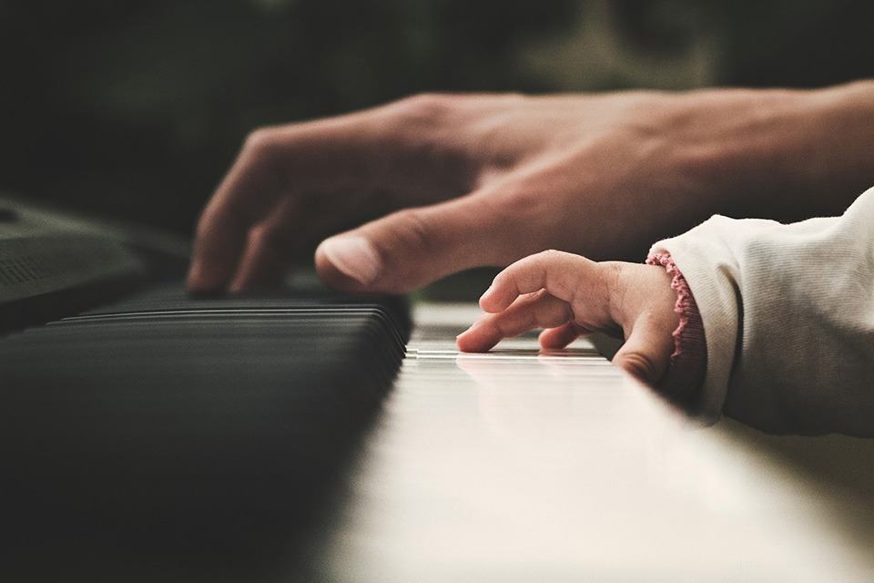 A close-up shot of a young child's hand, trying to play on a piano's white keys, with an adult hand in the background, instructing them.